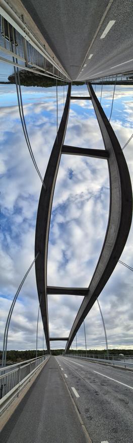 Panoramabild einer Brücke, aufgenommen mit einem Schwenk von der Straße links kopfüber den Brückenbögen folgend zur Straße rechts. Damit sieht man die Straße unten und oben, verbunden mit den (leicht schwankend aufgenommenen) Brückenbögen. Der bewölkte Himmel ist hinter den Bögen zu sehen.