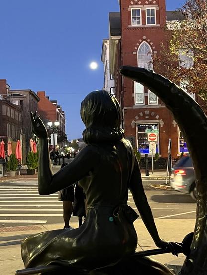 A small city at night scene. Foreground: a rear view of a bronze statue of Elizabeth Montgomery as Samantha from Bewitched. She's sitting side-saddle on a broom which is resting across a crescent moon. Directly above her head in the sky is a very bright full moon. Middle distance: a sidewalk and intersection. Background, multiple 19th century brick buildings on either side of a pedestrian-only street. To the right, is the four-story Daniel Low building, a historic jewelry company.