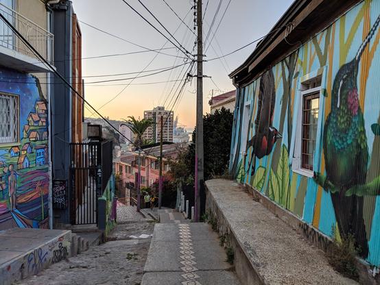 La imagen muestra una vista de una escalera en Valparaíso, Chile. Las paredes a ambos lados están cubiertas de murales que representan casas coloridas en una ladera y un colibrí de garganta roja. La escalera serpentea entre vallas de patios y edificios pintados de colores vivos. A lo lejos, se aprecian las siluetas del Cerro Concepción y el puerto de Valparaíso.