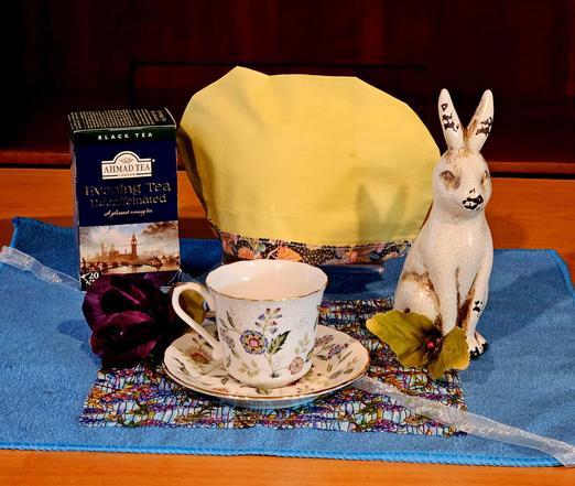 Tea tableau on blue French terry with a Tiffany panel and gauze ribbon. There is a rustic white ceramic rabbit casting a shadow of its bunny ears upon a yellow tea cozy. There are a white cup and saucer with a flower design and a dark maroon rose. The tea is Evening Tea Decaffeinated, a black tea blend from Ahmad.