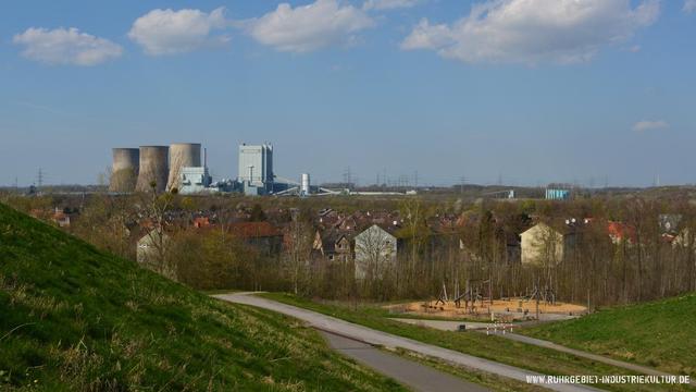Blick von einer Halde auf Wohnsiedlung mit Kohlekraftwerk im Hintergrund. Im Vordergrund grüne Böschung mit Weg und Spielplatz, dahinter Wohnhäuser zwischen kahlen Bäumen, am Horizont Kraftwerk mit Kühltürmen und Strommasten unter blauem Himmel.
