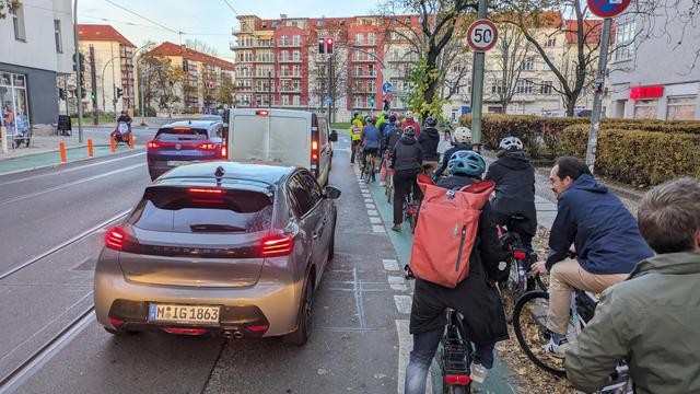 Viele Fahrradfahrende warten an der roten Ampel auf dem viel zu schmalen Fahrradstreifen während links von ihnen zweispurig die Autos stehen.