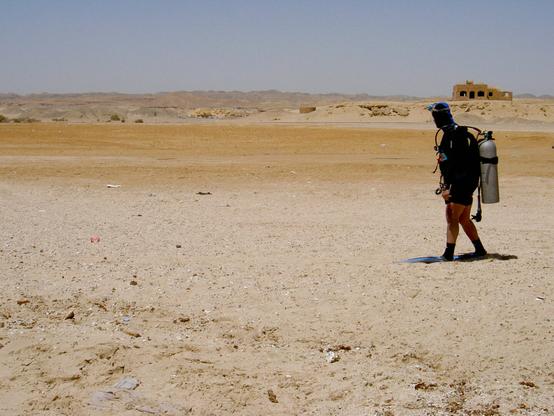 A person dressed in scuba gear walks across a sandy, arid landscape. Behind them is a distant structure, and the terrain is mostly barren with scattered debris.