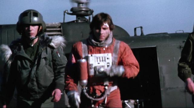 Richy Müller als Techniker Billy Hayes in orangefarbenem Raumanzug mit Helm und Ausrüstung vor einem Militär-Hubschrauber, flankiert von zwei Personen in dunkler Militärkleidung mit Helmen.