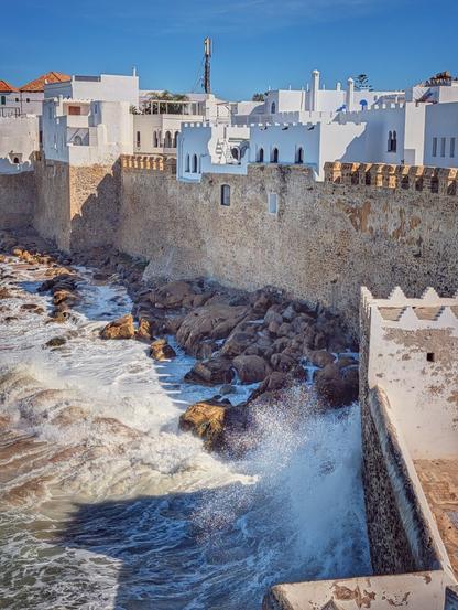 A vertical, sunny view of a rough Atlantic ocean crashing against the stone seawall and rocky base of a white-washed Moroccan town. The ancient, textured stone wall runs across the frame, supporting bright white buildings with minimal windows visible above. Foaming white waves surge over dark boulders, with a dramatic spray of water sparkling in the bright sunlight near the centre-right. The scene conveys a sense of enduring history and the powerful, dynamic meeting of sea and architecture.