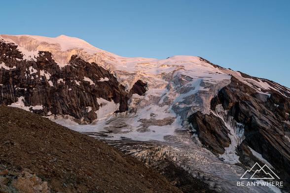 Soft morning light touching the snow-covered peaks and glaciers of a rugged alpine mountain range, the first warm tones of sunrise highlighting the textures of ice and rock.