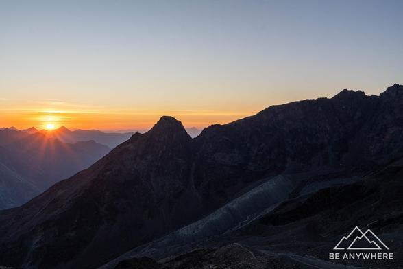 Sunrise over dark mountain silhouettes with the sun’s rays spreading across the horizon, casting a golden glow over the peaceful alpine landscape.
