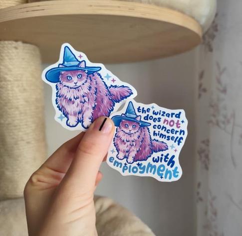 hand holding up two stickers of a kitty with a wizard hat, once without quote and once with "the wizard does not concern himself with employment"