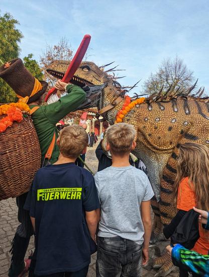 A lively scene from a Halloween event at Leipzig Zoo, featuring a strikingly realistic dinosaur costume with detailed feathers along its back. The dinosaur, possibly inspired by a feathered theropod, is being playfully tended to by a handler dressed in a green outfit and top hat. The handler is brushing the dinosaur’s teeth with an oversized red toothbrush, adding a fun and imaginative element to the display.

In the foreground, three children are captivated by the spectacle. One child wears a dark blue T-shirt with "Oberhainer Jugend Feuerwehr" printed on it, while the others are dressed in grey and orange tops. The event takes place outdoors, with trees and a clear sky in the background.