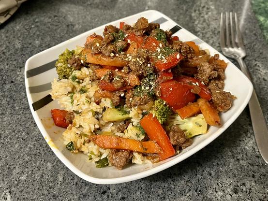 A square bowl of food, loaded with rice and topped with a medley of meat carrot and red capsicum. To the right is a fork