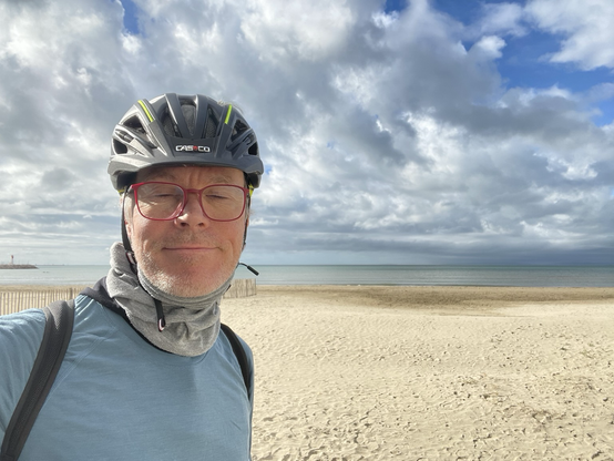 Mann mit Fahrradhelm vor einem Sandstrand. Im Hintergrund das Mittelmeer.