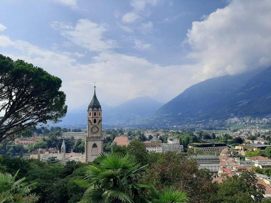 Blick auf Meran von oben, man sieht im Vorderbrund Bäume und Palmen, dann den ort mit einem großen Kirchturm im Zentrum des Bildes, dahinter Berge aufragen, obendrüber Wolken und blauer Himmel.
