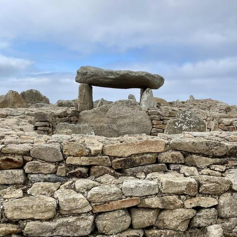 Im Vordergrund zu einer Mauer aufgeschichtete Steine. Mittig ein Dolmen ("Steintisch"), bestehend aus mehreren großen, aufrecht stehenden Tragsteinen. Auf den Tragsteinen liegt eine riesige Deckplatte.