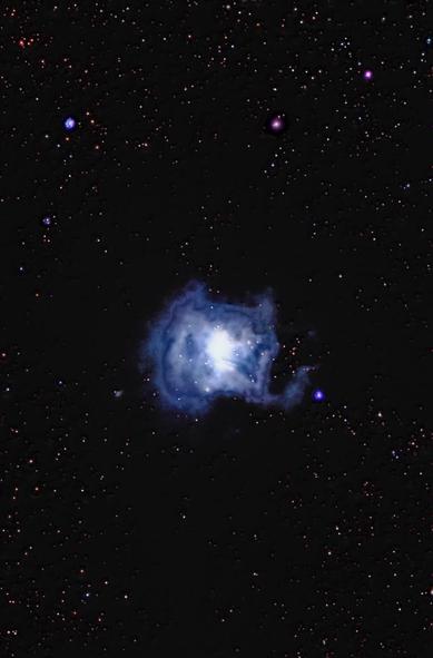 Jasna mgławica znajduje się w centrum obrazu, otoczona ciemnym niebem wypełnionym licznymi gwiazdami. Mgławica wykazuje świetliste, niebieskawo-białe jądro z cienkimi, przypominającymi chmury formacjami.