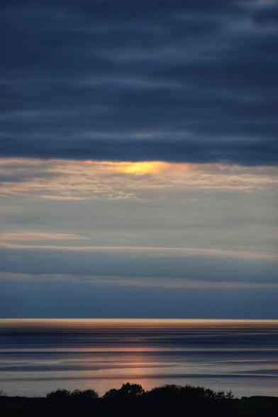 A serene coastal scene during twilight, with the sky transitioning from deep blues to soft oranges and yellows near the horizon. Thin, wispy clouds stretch across the sky, reflecting the fading light of the sun. The calm sea below mirrors the colours of the sky, creating a peaceful gradient of muted blues and golds. The silhouette of trees or shrubs is faintly visible along the bottom edge, adding depth to the scene. The overall atmosphere is tranquil and contemplative, capturing the quiet beauty of nature at the end of the day.