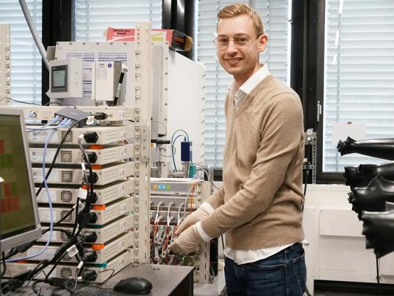 Researcher Fabian Apfelbeck, wearing glasses and a beige sweater, works in a laboratory with electronic measurement equipment and monitoring devices.