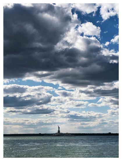 Photo of the waters of New York Bay under a sky heavy with low, shadowy clouds. On the horizon is Liberty Island, where the tiny figure of the Statue of Liberty stands silhouetted against an area of brighter sky. Beyond the island is the low New Jersey coastline.