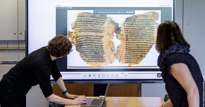Zwei Personen betrachten auf einem großen Bildschirm die Abbildung eines antiken Papyrus mit griechischem Text; eine Person arbeitet an einem Laptop.