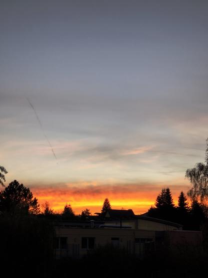 Auf dem Foto sieht man im Vordergrund ein paar Dächer und Bäume. Darüber geht gerade die Sonne unter und schafft ein Spektrum an roten Farbtönen. Im Hintergrund sind ein paar dunkle Schleierwolken zu sehen.