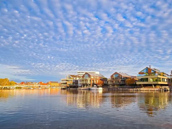 Blue water can be seen in the lower part of the picture. Above it is a blue sky with lots of small white clouds. They surely have a meteorological name, but I don't know what it is. Houses can be seen between the sky and the water. They have terraces facing the water. A little further on, the water bends. There are more houses there in the sun and a bridge connecting the two banks. It is pretty.