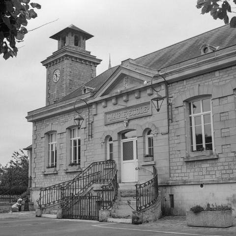 Black and white photo of a french town hall with a small clock tower. An ornate staircase leads to the entrance under a coat of arms and a large sign "Mairie Ecole"