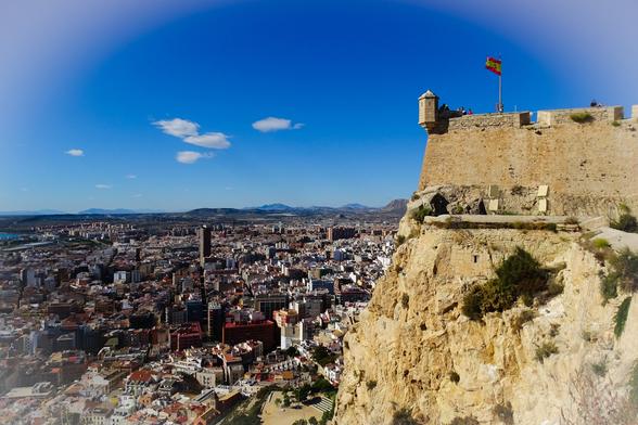 Näkymä linnoitukselta. Oikealla Benacantil-kukkulan jyrkkää rinnettä ja linnoituksen muuri, vasemmalla kaukana alhaalla Alicanten kaupunki. Sininen taivas ja auringonpaiste.
