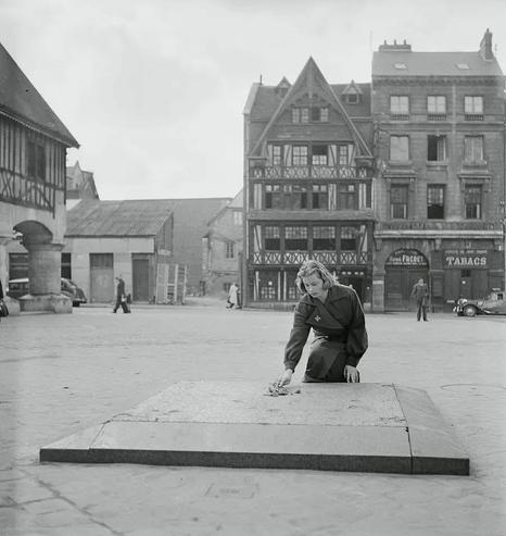 Black white photo of Ingrid Bergman putting a small bouquet on a platform in the middle of a square. Ancient looking buildings in the back. some people are in the streets, at the square.
