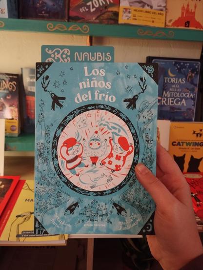 Fotografía del libro: en tonos blancos, negros, azules y naranjas hay una composición que remite a frío. Aparecen sus protagonistas en el centro dibujados dentro de un círculo blanco, mientras que el resto de la portada tiene fondo azul y pequeños detalles y dibujos de naturaleza, agua y magia.