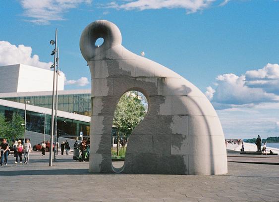 The first photo in the diptych, showing a large and abstract stone sculpture in an open space outside the library, with the opera building in the background. People are walking by, sightseeing and enjoying the weather.