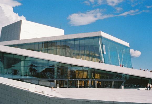The second photo in the diptych, showing the Oslo Opera House with its glass walls and snow-white, walkable roof against a bright blue sky with puffy, white clouds.