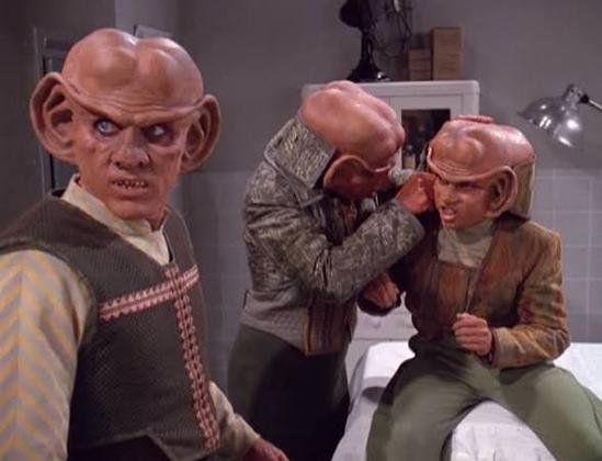 Quark, Nog, and Rom in a 1940s military facility health unit with Rom inspecting the inside of Nog’s ears