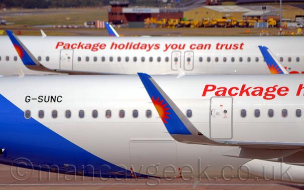 Close-up of the up-turned wingtips of a white jet airliner parked facing to the right, with another, nearly identical plane in the background.
The plane is mostly whit, with a blue lower rear fuselage, reaching up towards the tail, , with the black registration "G-SUNC" on the upper rear.
There is red text starting with the word "Package" on the upper fuselage towards the right - the plane in the background has the full text "Package holidays you can trust" running along the fuselage.
The up-turned wing-tips are blue, with a yellow, orange, and red sunburst coming from the rear.
In the distance, yellow airport vehicle sare clustered around a large yellow shed and a pair of brown brick buildings.