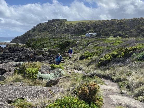 Two hikers walk along a coastal path surrounded by green bushes, with hills and a bit of ocean visible behind them under partly cloudy skies.