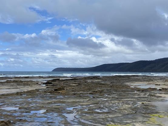 A view across a beach with hills visible in the distance under partly cloudy skies.