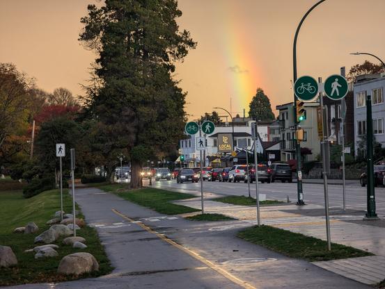 A photo taken from the side of a separated bike path on the edge of a park. Beside the bike path is a sidewalk and then the street. There are some 2 and 3 storey buildings on the far side of the road. There are cars on the road, in both directions, but not heavy traffic. There is a big tree in the middle distance. The sky is orange-grey except for a bright rainbow on the right.