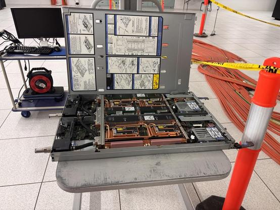 This is what is inside the slots on the rack. Very efficient! We’re looking at an open case containing drives and circuit boards with very shiny copper pipes for liquid cooling. It’s sitting on a little table next to an orange bollard with “do not cross” tape hanging off it.