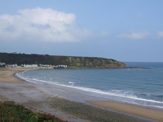 A sandy beach curves gently along a calm bay, with small waves lapping at the shore and scattered yellow buoys floating in the water. A cluster of white houses and low buildings sits near the shoreline, backed by dark green cliffs with patches of exposed rock. The headland extends into the sea, sheltering the bay, and the open water stretches to the horizon under a mostly clear blue sky with some scattered clouds. The foreground includes wet sand and small areas of pebbles, while the tide appears to be receding.