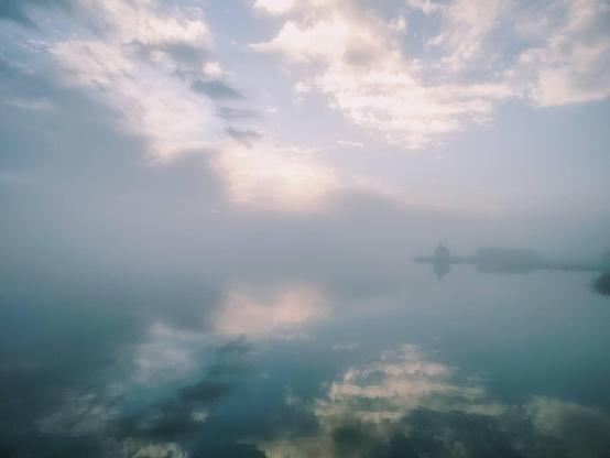 Photo of fog over the water but also with the sky and clouds reflecting in the still sea. In the distance beyond the fog a barely visible little chapel on a pier.
