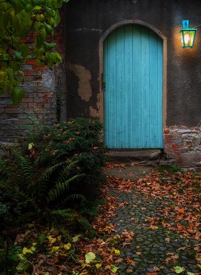 A charming, rustic scene featuring a weathered building with a striking turquoise door set into a dark, textured wall. The door is framed by an arch of aged stonework, adding to the vintage atmosphere. To the right of the door, a matching turquoise lantern hangs, casting a warm and inviting glow. The ground is paved with cobblestones, scattered with autumn leaves in shades of green, brown, orange, and yellow. Lush green ferns and shrubs grow along the left side, softening the rugged appearance of the brickwork and enhancing the picturesque, timeless quality of the setting.