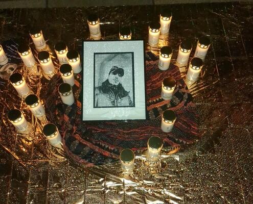 Schwarz-weiß Foto eines jungen Mannes mit Baseballkappe, in einem Rahmen, umgeben von brennenden Grablichtern