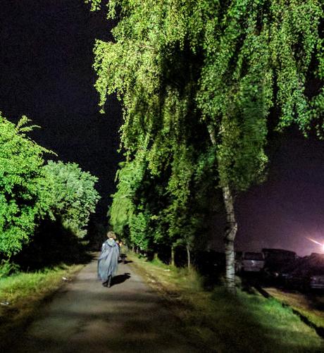 Ein landwirtschaftlicher Weg, mit Birken rechts und hohem Gebüsch links begrünt. Sehr helle Strahler heben das Grün sehr kontrastreich gegen die mitternächtliche Dunkelheit ab. In der Mitte des Weges ist sehr weit vor eine Frau mit lockigem Haar und langem grauen Regencape zu erkennen, sich vom Festivalgelände entfernend.