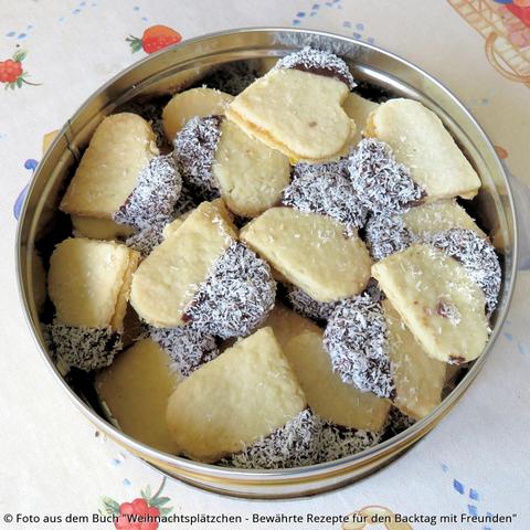 Auf dem Tisch eine Blechdoese mit vielen gefüllten Kokosherzen. Die Kese sind mit Marmelade zusammengesetzt. Eine Herzhälfte hat einen Schokoüberzug und ist mit Kokos bestreut.