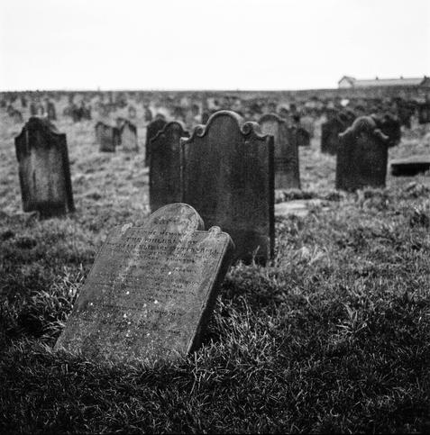 Black and white photo of a graveyard. There are graves dotted across the field - in the foreground a stone has fallen and lays tilted so the inscription is hard to make out.