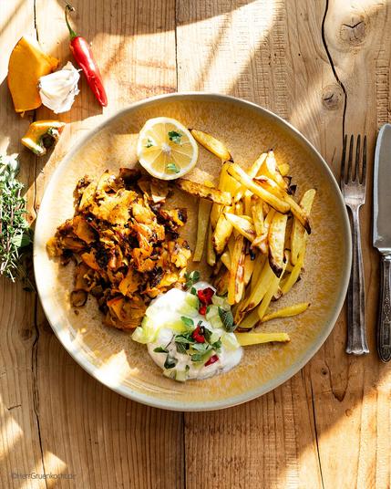 Holzuntergrund. Ein runder gelber Teller. Darauf: Kürbis-Kebap mit eigener Gewürzmischung, Pommes-Frites und einem Joghurt-Gurken-Topping