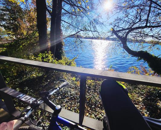 Blick auf einen blauen See zwischen Herbstlaubbäume hindurch, gegen Sonnenlicht im wolkenfreien Himmel. Im Bildvordergrund unten ein blauschwarzes Fahrrad und ein Bein in Radklamotten an einem Geländer abgestützt. Unten links hält eine Hand den Lenker.