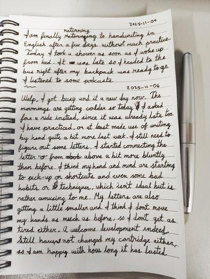 A notebook with a pen by its side, the following text in cursive:

I am finally returning to handwriting in English after a few days without much practice. Today I took a shower as soon as I woke up from bed. It was late so I headed to the bus right after my backpack was ready to go. I listened to some podcasts...

Welp I got busy and it's a new day now. The mornings are getting colder so today I asked for a ride instead, since it was already late too. I have practiced, or at least made use of writing by hand quite a bit more last week. I still need to figure out some letters. I started connecting the letter "o" from above a bit more bluntly than before, I think my hand and mind are starting to pick-up on shortcuts and even some bad habits or techniques, which isn't ideal but is rather amusing to me. My letters are also getting a little smaller and I think I don't move my hands as much as before, so I don't get as tired either. A welcome development indeed.

Still have not hanged my cartridge either so I am happy with how long it has lasted