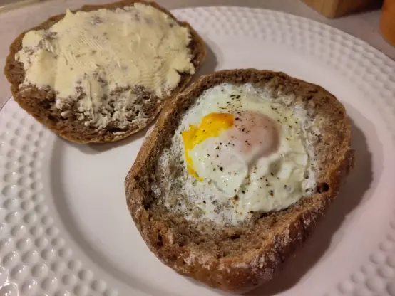 Eine Brötchenhälfte wurde in der Mikrowelle mit einem Ei gegart. Daneben liegt die übrige Brötchenhälfte bestrichen mit Margarine und bestreut mit Salz.