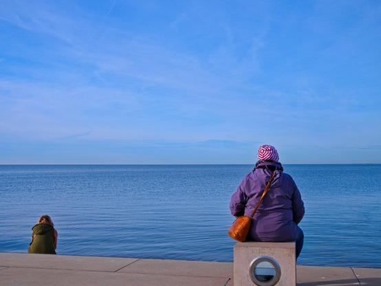 By the sea. Two women are sitting, some distance apart, on a concrete block and a little further away on a step. They have their backs to me and are looking out at the calm North Sea. The water is blue, as is the sky. A very peaceful picture.