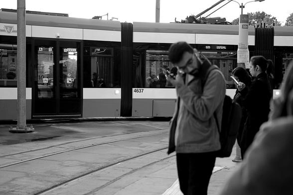 The outdoor platform. Across the background of the image, side to side, a streetcar makes its turn to leave the station. Right of centre midground is a bearded man, his head turned toward the camera and tilted down. He is his left hand is touching the right side of his head. At far right are other waiting passengers.