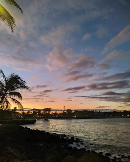 sunset in a bay of a tropical island. the street lights are already litten and the ocean is very calm. on the side of the bay there are some palm trees and there are some scattered clouds in the sky
